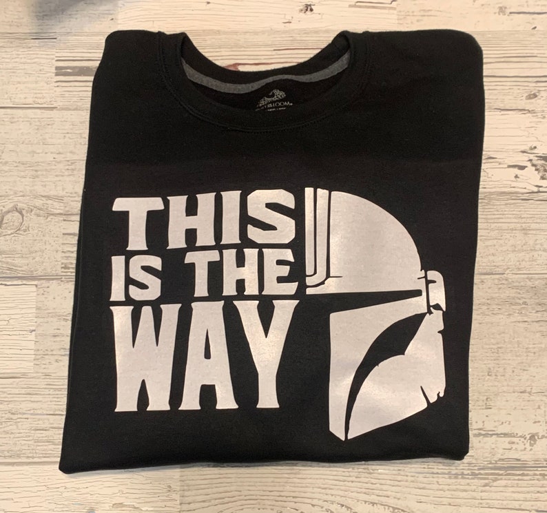 Puede incluir: Camiseta negra con un estampado gr&aacute;fico blanco de un casco de Mandaloriano y el texto "This is the way".