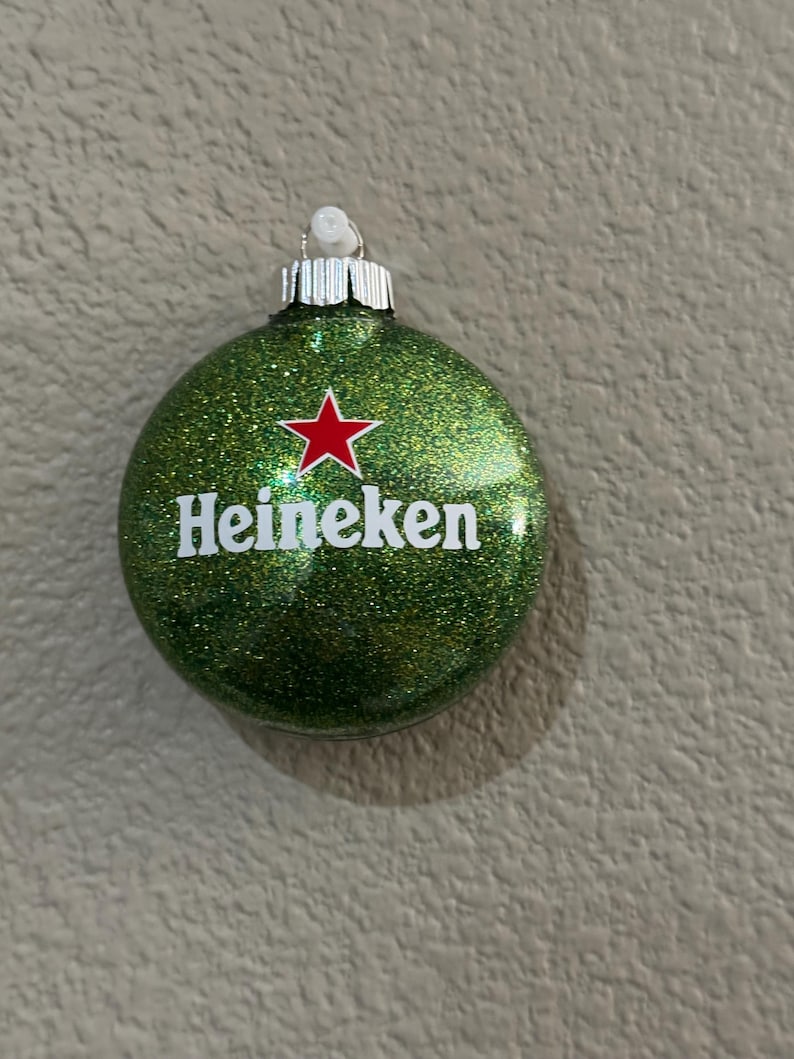 Puede incluir: Adorno navide&ntilde;o verde brillante con el logo de Heineken. El adorno tiene una tapa plateada y una estrella roja encima de la palabra "Heineken". El adorno es redondo y tiene una textura brillante.