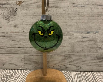 Puede incluir: Adorno navideño de vidrio verde con acabado brillante y una cara de Grinch con ojos amarillos.