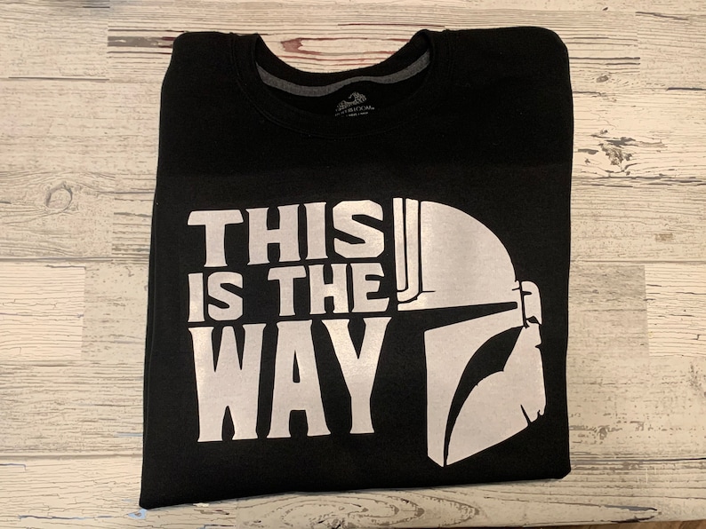 Puede incluir: Sudadera negra con un estampado blanco de un casco de Mandaloriano y el texto "This is the Way".