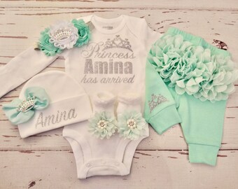May include: A baby girl outfit set in white and mint green. The set includes a bodysuit, pants, a hat, a headband, and booties. The bodysuit has a silver glitter design that says "Princess Amina has arrived". The hat and pants have a silver glitter design that says "Amina". The headband has a white flower and a silver crown.