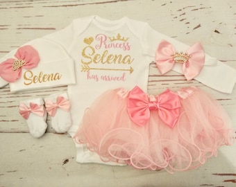 Peut inclure: Un ensemble de vêtements pour bébé fille rose et or comprenant un body blanc, une jupe tutu rose, un bandeau à nœud rose, un chapeau à nœud rose et des chaussons rose et blanc. Le body est orné d'un texte en paillettes dorées qui dit "Princess Selena has arrived". Le chapeau est orné d'un texte en paillettes dorées qui dit "Selena".