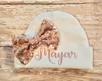 May include: A white baby beanie with a large rose gold sequin bow. The name "Mayar" is printed in a matching rose gold script across the front of the hat. The hat is made of soft material.