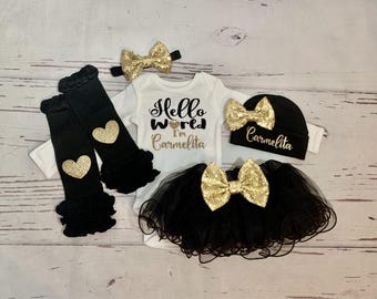Peut inclure: Un body blanc à manches longues avec le texte noir et doré Hello World I'm Carmelita, un tutu en tulle noir avec un nœud à paillettes dorées, des jambières noires à cœurs dorés, un bonnet noir avec texte doré et un bandeau à paillettes.