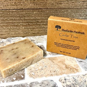 May include: A brown cardboard box with the text "SheaGarden Handmade Cedar Tree Handmade Soap Bar" and the text "Made with raw unrefined fats and oils. Naturally lathering and skin cleansing with a moisturizing smooth feel Net.WT 3.5 OZ". A bar of soap is sitting on a tiled surface.
