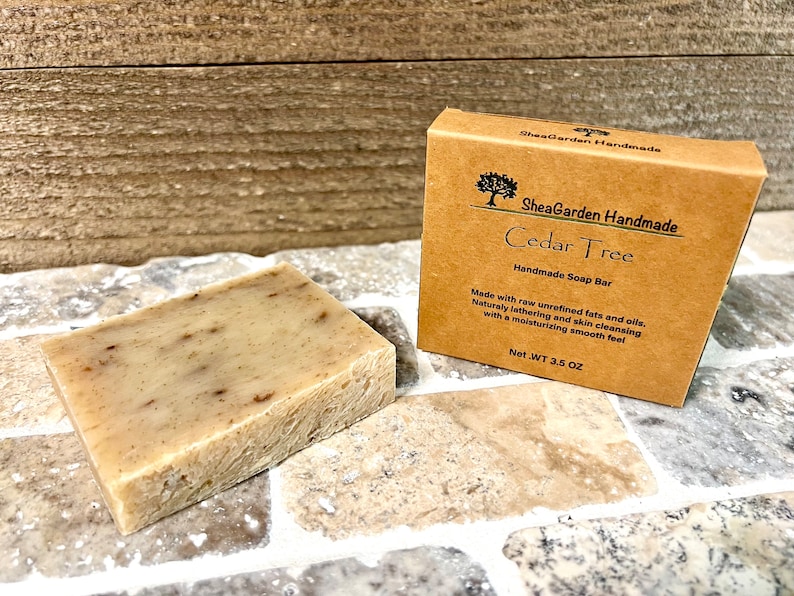 May include: A brown cardboard box with the text "SheaGarden Handmade Cedar Tree Handmade Soap Bar" and the text "Made with raw unrefined fats and oils. Naturally lathering and skin cleansing with a moisturizing smooth feel Net.WT 3.5 OZ". A bar of soap is sitting on a tiled surface.
