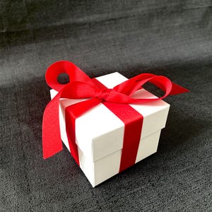 K&ouml;nnte beinhalten: Eine kleine, quadratische wei&szlig;e Geschenkbox mit einem leuchtend roten Satinband, das zu einer Schleife gebunden ist. Das Band umschlingt die Box und sorgt f&uuml;r eine festliche und elegante Pr&auml;sentation. Die Box steht auf einer dunkelgrauen strukturierten Oberfl&auml;che.