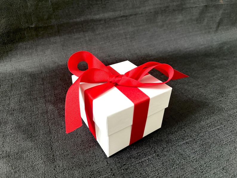 K&ouml;nnte beinhalten: Eine kleine, quadratische wei&szlig;e Geschenkbox mit einem leuchtend roten Satinband, das zu einer Schleife gebunden ist. Das Band umschlingt die Box und sorgt f&uuml;r eine festliche und elegante Pr&auml;sentation. Die Box steht auf einer dunkelgrauen strukturierten Oberfl&auml;che.