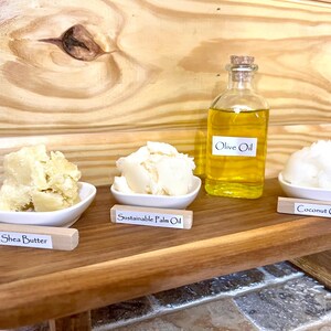 May include: A wooden tray with four small bowls containing shea butter, sustainable palm oil, coconut oil, and a bottle of olive oil. Each bowl has a small wooden sign with the name of the oil or butter.