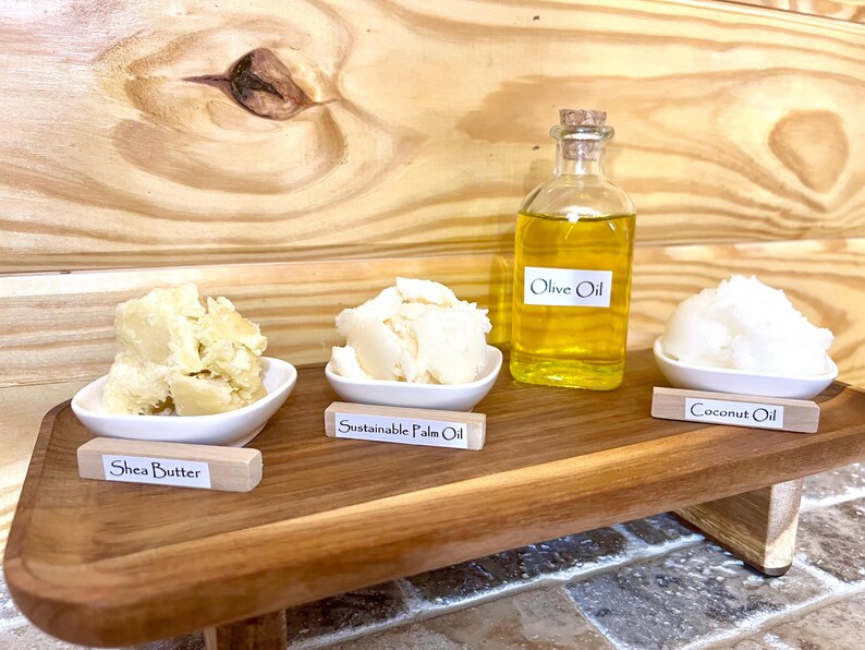 May include: A wooden tray with four small bowls containing shea butter, sustainable palm oil, coconut oil, and a bottle of olive oil. Each bowl has a small wooden sign with the name of the oil or butter.