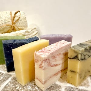 K&ouml;nnte beinhalten: Eine Auswahl handgemachter Seifen in verschiedenen Farben und Mustern. Die Seifen umfassen gr&uuml;ne, marineblaue, gelbe, rosa und marmorierte Designs. Ein Stapel wei&szlig;er Waschlappen und eine kleine verpackte Seife befinden sich im Hintergrund.