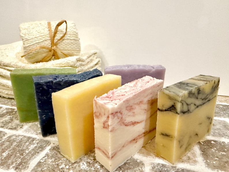 K&ouml;nnte beinhalten: Eine Auswahl handgemachter Seifen in verschiedenen Farben und Mustern. Die Seifen umfassen gr&uuml;ne, marineblaue, gelbe, rosa und marmorierte Designs. Ein Stapel wei&szlig;er Waschlappen und eine kleine verpackte Seife befinden sich im Hintergrund.