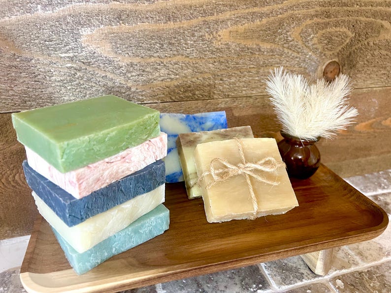 K&ouml;nnte beinhalten: Eine Auswahl handgemachter Seifen in verschiedenen Farben, darunter Gr&uuml;n, Rosa, Blau und Beige, auf einem Holztablett pr&auml;sentiert. Einige Seifen sind mit Bindfaden gebunden. Eine kleine braune Vase mit wei&szlig;en Zierfedern ist ebenfalls vorhanden.