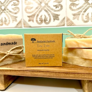Könnte beinhalten: Zwei handgemachte Seifenstücke, eines mit der Aufschrift "Handmade" und das andere mit der Aufschrift "SheaGarden Handmade Tea Tree Handmade Soap Bar". Die Seifenstücke sind mit Schnur umwickelt und liegen auf einem Holzregal.