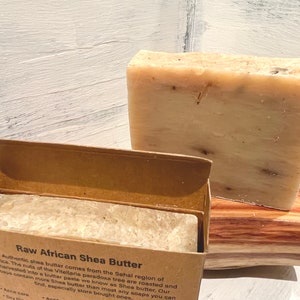 May include: Two bars of raw African shea butter soap. One bar is in a brown cardboard box with the text "Raw African Shea Butter" and a description of the product. The other bar is on a wooden surface.