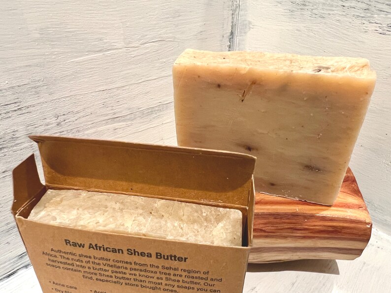 May include: Two bars of raw African shea butter soap. One bar is in a brown cardboard box with the text "Raw African Shea Butter" and a description of the product. The other bar is on a wooden surface.