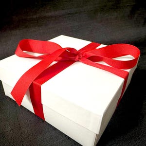 K&ouml;nnte beinhalten: Eine wei&szlig;e Geschenkbox, gebunden mit einem leuchtend roten Band. Das Band umschlie&szlig;t die Box und ist oben zu einer Schleife gebunden. Die Box steht auf einer dunklen, strukturierten Oberfl&auml;che.