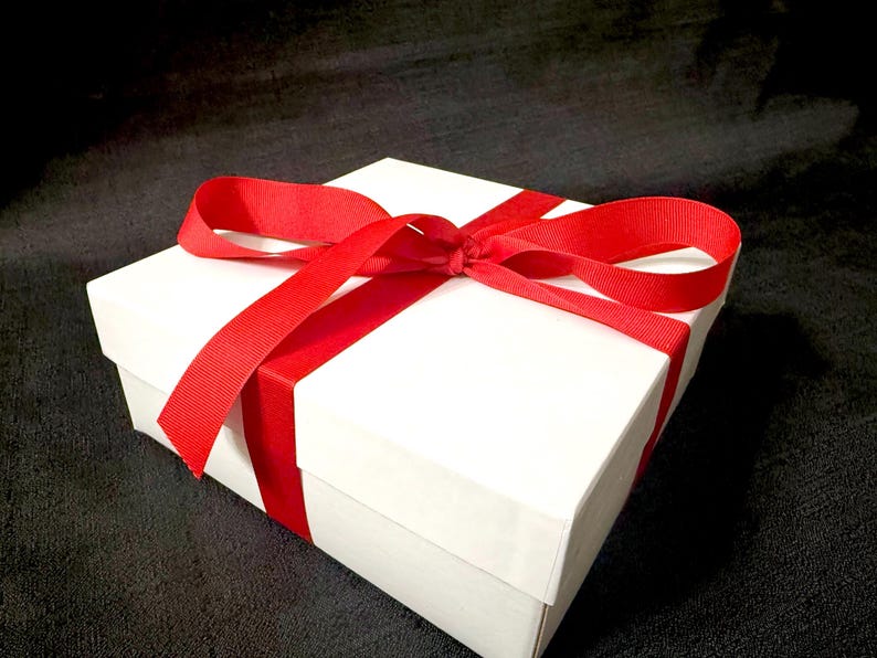 K&ouml;nnte beinhalten: Eine wei&szlig;e Geschenkbox, gebunden mit einem leuchtend roten Band. Das Band umschlie&szlig;t die Box und ist oben zu einer Schleife gebunden. Die Box steht auf einer dunklen, strukturierten Oberfl&auml;che.