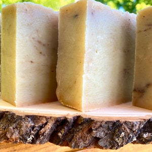 May include: Four bars of handmade soap, each with a light brown and white marbled appearance, are displayed on a wooden slice. The soap bars are rectangular and have a smooth, polished surface.