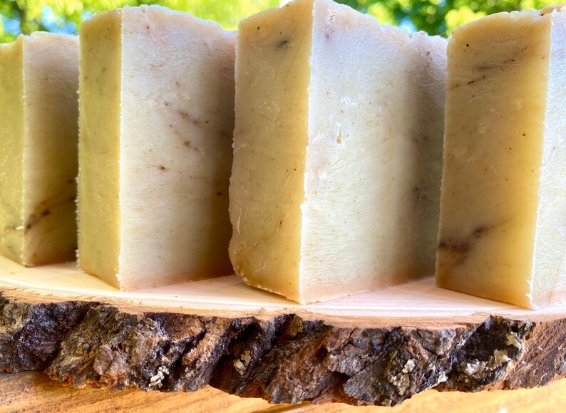 May include: Four bars of handmade soap, each with a light brown and white marbled appearance, are displayed on a wooden slice. The soap bars are rectangular and have a smooth, polished surface.