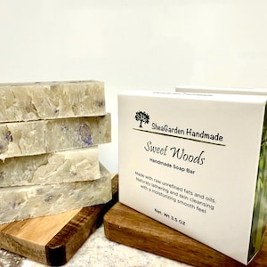 May include: Five bars of handmade soap stacked on a wooden surface. The soap is light beige with flecks of purple. The soap is packaged in a white box with green accents and the text "SheaGarden Handmade Sweet Woods Handmade Soap Bar Made with raw unrefined fats and oils. Naturally lathering and skin cleansing with a moisturizing smooth feel Net. Wt 3.5 OZ"