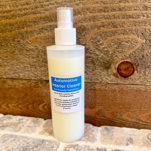 May include: A clear plastic bottle with a white pump top and a blue label that reads "Automotive Interior Cleaner - Eco-Friendly Ingredients". The bottle is filled with a white liquid and is sitting on a wooden surface.