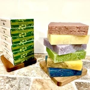 以下が含まれることがあります： 紫、緑、青など、さまざまな色の手作り石鹸が木製の台に並んでいます。一部の石鹸は、緑色のラベルが付いた白い箱に梱包されています。この画像は、さまざまな天然石鹸を紹介しています。