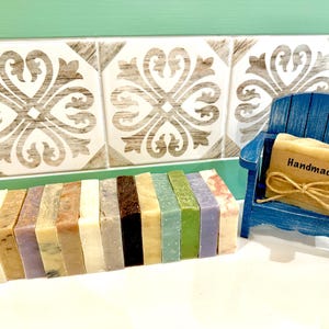 K&ouml;nnte beinhalten: Eine Sammlung handgemachter Seifen in verschiedenen Farben und Texturen. Ein kleiner blauer Holzstuhl h&auml;lt ein Seifenst&uuml;ck mit einem "Handmade"-Etikett, das mit Bindfaden gebunden ist. Die Seifen sind auf einer wei&szlig;en Oberfl&auml;che angeordnet.