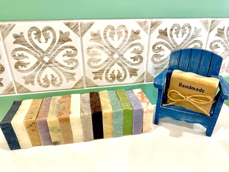 K&ouml;nnte beinhalten: Eine Sammlung handgemachter Seifen in verschiedenen Farben und Texturen. Ein kleiner blauer Holzstuhl h&auml;lt ein Seifenst&uuml;ck mit einem "Handmade"-Etikett, das mit Bindfaden gebunden ist. Die Seifen sind auf einer wei&szlig;en Oberfl&auml;che angeordnet.