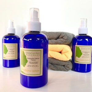May include: Three blue bottles of eucalyptus lemon automotive interior cleaner with a white spray nozzle. The bottles are labelled "SheaGarden Handmade" and "Automotive Interior Cleaner". The bottles are sitting on a white surface with two folded towels behind them.
