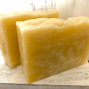 May include: Two rectangular bars of handmade soap. The soap is a pale yellow color with a marbled appearance. The bars are sitting on a white, distressed wooden surface. The soap has a slightly rough, textured top.