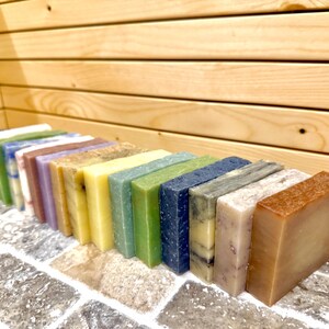 K&ouml;nnte beinhalten: Eine Sammlung rechteckiger handgemachter Seifen in verschiedenen Farben, darunter Blau, Gr&uuml;n, Gelb und Braun. Die Seifen sind in einer Reihe auf einer Ziegeloberfl&auml;che mit einem h&ouml;lzernen Hintergrund angeordnet.