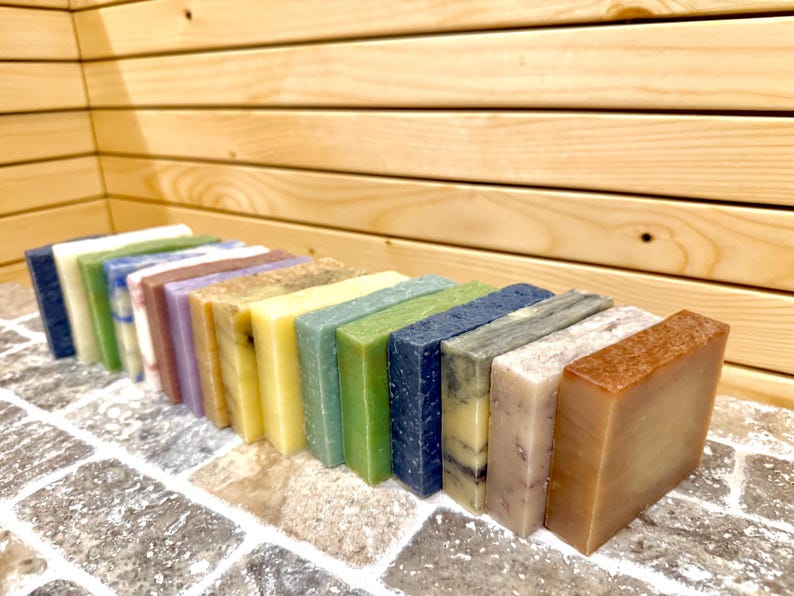 K&ouml;nnte beinhalten: Eine Sammlung rechteckiger handgemachter Seifen in verschiedenen Farben, darunter Blau, Gr&uuml;n, Gelb und Braun. Die Seifen sind in einer Reihe auf einer Ziegeloberfl&auml;che mit einem h&ouml;lzernen Hintergrund angeordnet.
