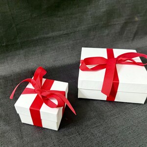 K&ouml;nnte beinhalten: Zwei wei&szlig;e Geschenkboxen mit roten Satinb&auml;ndern und Schleifen. Die quadratischen Boxen sind unterschiedlich gro&szlig;. Die B&auml;nder sind um die Boxen gewickelt und oben zu Schleifen gebunden. Auf einer dunkelgrauen Oberfl&auml;che.