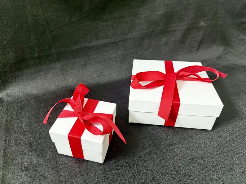 K&ouml;nnte beinhalten: Zwei wei&szlig;e Geschenkboxen mit roten Satinb&auml;ndern und Schleifen. Die quadratischen Boxen sind unterschiedlich gro&szlig;. Die B&auml;nder sind um die Boxen gewickelt und oben zu Schleifen gebunden. Auf einer dunkelgrauen Oberfl&auml;che.