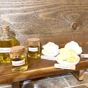 May include: A wooden tray with six glass bottles and jars containing various oils and butters. The labels on the bottles and jars read "Olive Oil", "Castor Oil", "Essential Oil", "Sustainable Palm Oil", "Shea Butter", and "Coconut Oil".