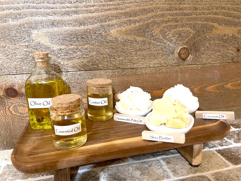 May include: A wooden tray with six glass bottles and jars containing various oils and butters. The labels on the bottles and jars read "Olive Oil", "Castor Oil", "Essential Oil", "Sustainable Palm Oil", "Shea Butter", and "Coconut Oil".
