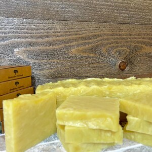 May include: A stack of four bars of handmade soap, with a yellow color and a slightly textured surface. The soap is sitting on a wooden surface, with a stack of boxes behind it. The boxes are brown with green and white text that reads "Botanical Handmades".