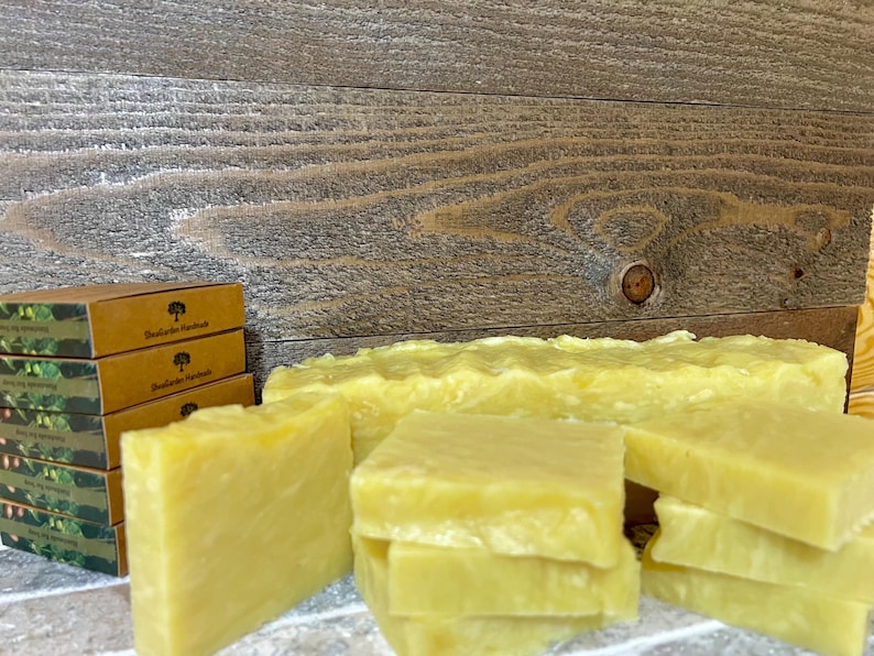 May include: A stack of four bars of handmade soap, with a yellow color and a slightly textured surface. The soap is sitting on a wooden surface, with a stack of boxes behind it. The boxes are brown with green and white text that reads "Botanical Handmades".