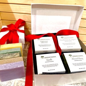 K&ouml;nnte beinhalten: Eine wei&szlig;e Geschenkbox mit rotem Band, neben einer wei&szlig;en Box mit roter Schleife. In der offenen Box befinden sich vier kleine wei&szlig;e Schachteln mit handgemachten Seifenst&uuml;cken und mehrere Seifenst&uuml;cke in verschiedenen Farben.