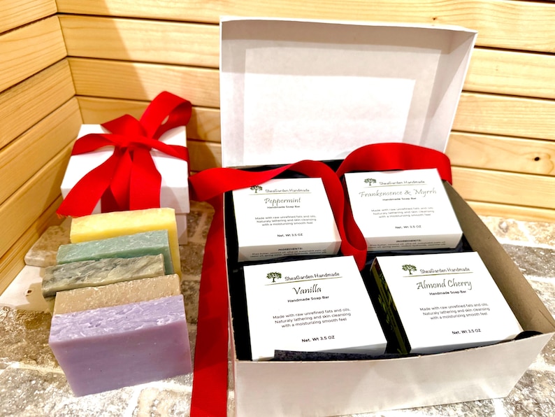 K&ouml;nnte beinhalten: Eine wei&szlig;e Geschenkbox mit rotem Band, neben einer wei&szlig;en Box mit roter Schleife. In der offenen Box befinden sich vier kleine wei&szlig;e Schachteln mit handgemachten Seifenst&uuml;cken und mehrere Seifenst&uuml;cke in verschiedenen Farben.
