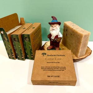 May include: A brown cardboard box with the text "SheaGarden Handmade Cedar Tree Handmade Soap Bar" and "Net.WT 3.5 OZ". The box is sitting on a wooden surface next to a bar of soap and a small gnome figurine.