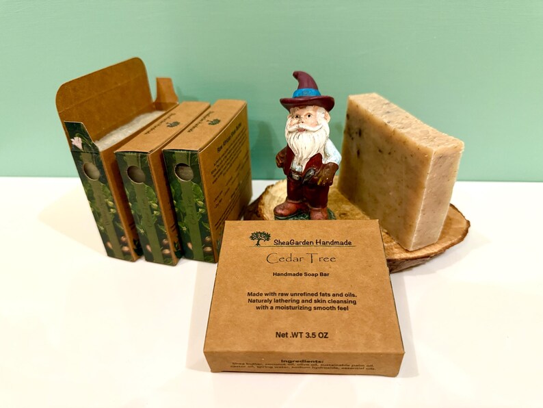 May include: A brown cardboard box with the text "SheaGarden Handmade Cedar Tree Handmade Soap Bar" and "Net.WT 3.5 OZ". The box is sitting on a wooden surface next to a bar of soap and a small gnome figurine.