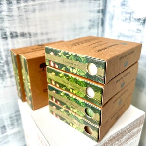 May include: A stack of four brown cardboard boxes with green and white labels. The labels feature a floral design and the text "Lemongrass", "Almond Cherry", "Green Tea", and "Lavender Rosemary".