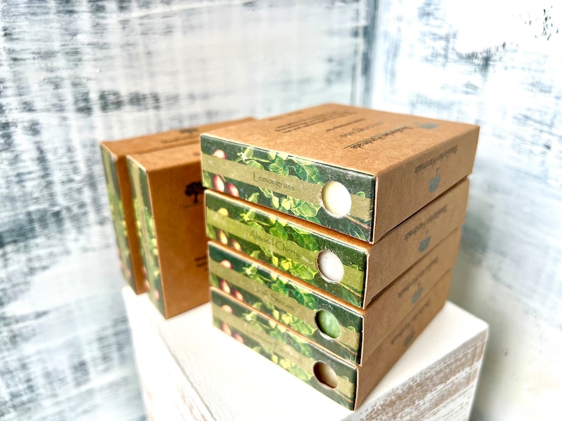 May include: A stack of four brown cardboard boxes with green and white labels. The labels feature a floral design and the text "Lemongrass", "Almond Cherry", "Green Tea", and "Lavender Rosemary".