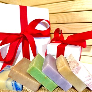 K&ouml;nnte beinhalten: Eine Auswahl an bunten, handgemachten Seifen in Blau-, Braun-, Gr&uuml;n-, Lila- und Beiget&ouml;nen. Die Seifen sind vor zwei wei&szlig;en Geschenkboxen mit roten B&auml;ndern angeordnet, was auf ein Geschenkset hindeutet.