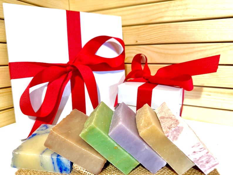 K&ouml;nnte beinhalten: Eine Auswahl an bunten, handgemachten Seifen in Blau-, Braun-, Gr&uuml;n-, Lila- und Beiget&ouml;nen. Die Seifen sind vor zwei wei&szlig;en Geschenkboxen mit roten B&auml;ndern angeordnet, was auf ein Geschenkset hindeutet.