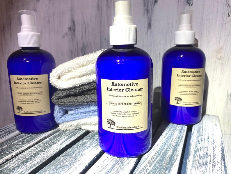 May include: Three blue spray bottles with white spray tops and labels that read "Automotive Interior Cleaner". The bottles are on a whitewashed wooden surface with a stack of towels behind the centre bottle.