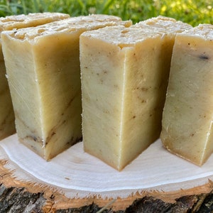 May include: Five bars of handmade soap, each with a light brown and white marbled appearance, are arranged in a row on a wooden surface.