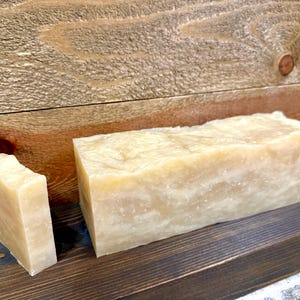 May include: A rectangular block of off-white soap with a marbled texture and a single smaller, thinner slice of the same soap standing upright next to it, both featuring a smooth, slightly uneven surface with subtle variations in color.